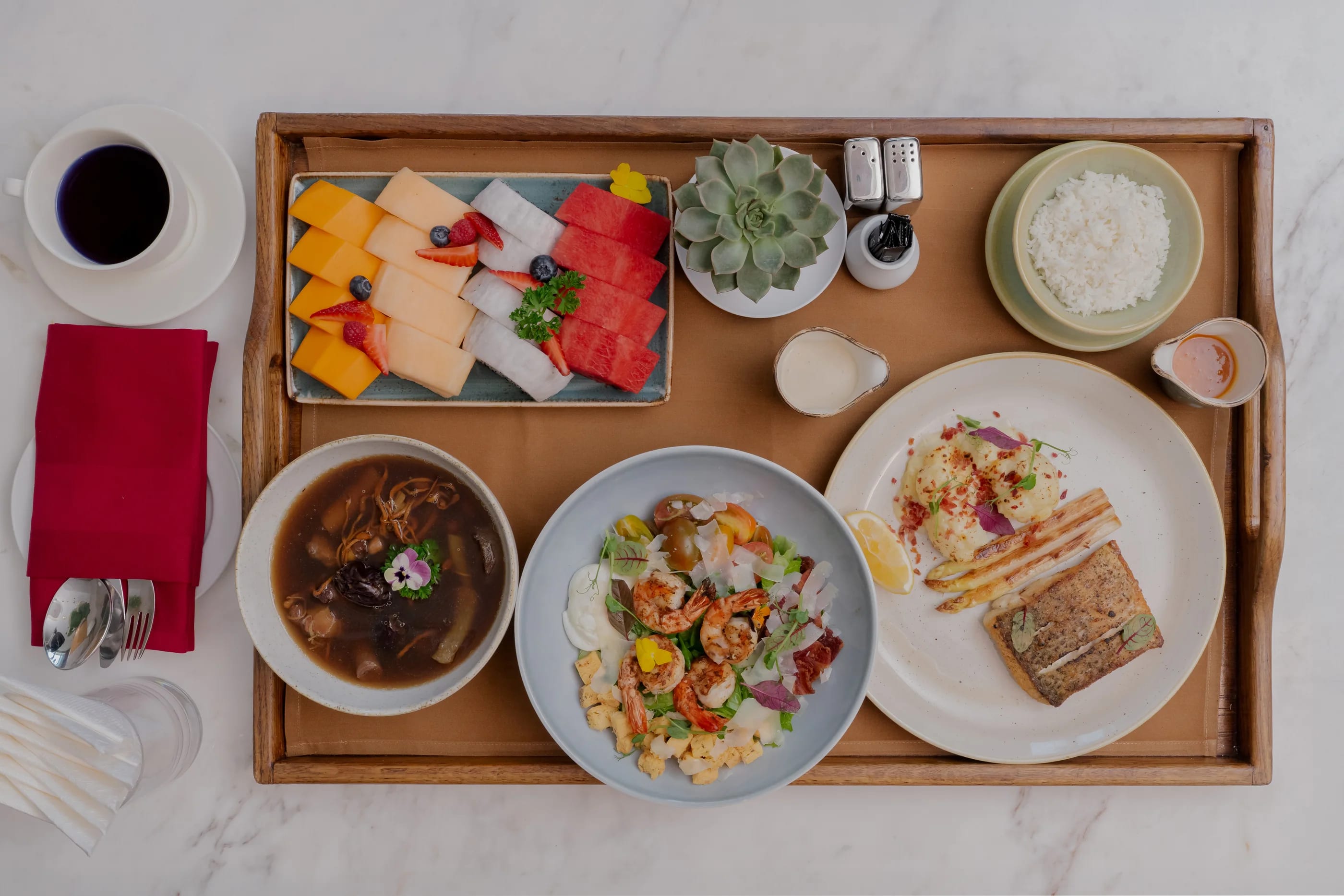A lunch tray seen from above: herbal broth, prawn salad, seared fish with asparagus, rice and fresh fruit