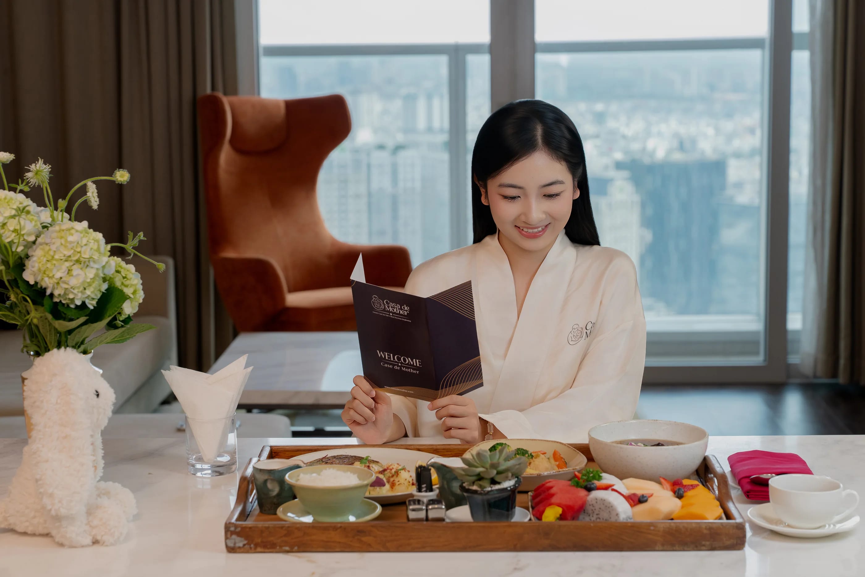 A mother in a Casa robe reading the welcome menu beside her breakfast tray, the city behind the window