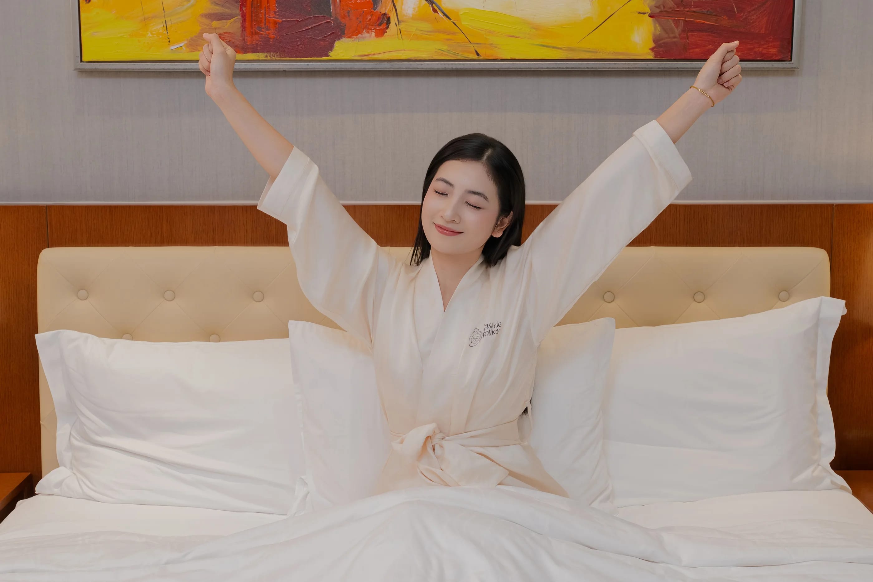 A mother in a Casa robe stretching awake on a white bed in her suite